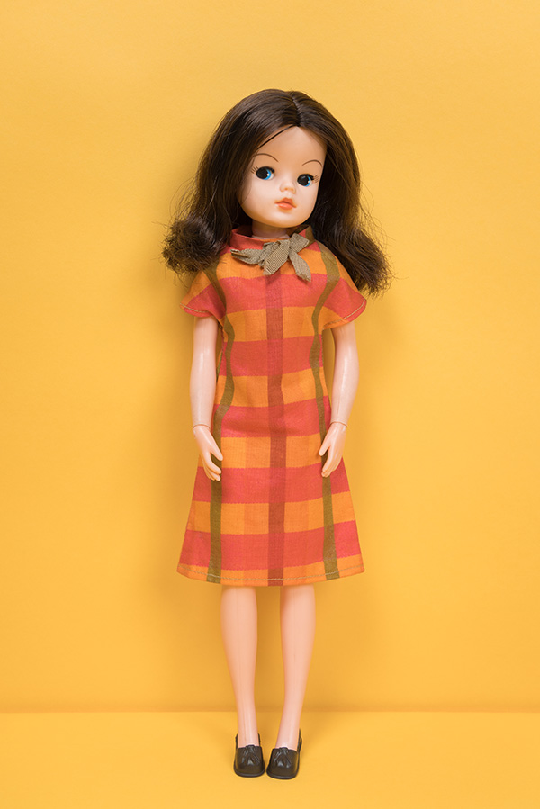 white doll with brunette hair wearing a short sleeved, knee length dress with orange, pink, and green plaid print