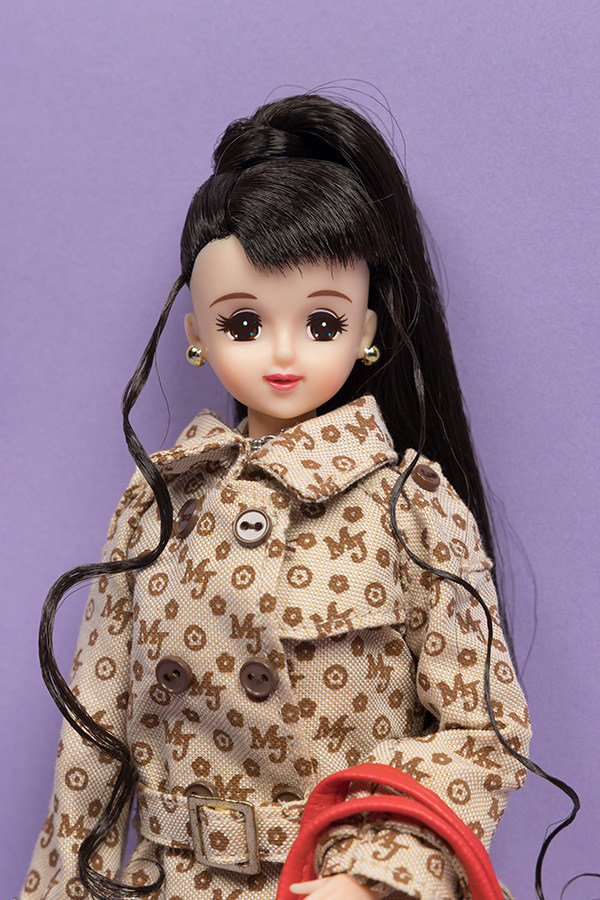 white doll with dark hair styled in a high ponytail and bangs, wearing a tan trenchcoat with a repeating print pattern of "MJ" and flowers