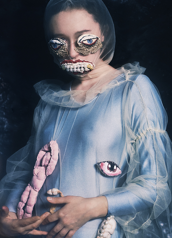photograph of seated woman wrapped in translucent tulle, eyes covered with embroidered eyes, pearls for teeth, and holding a pink knit doll