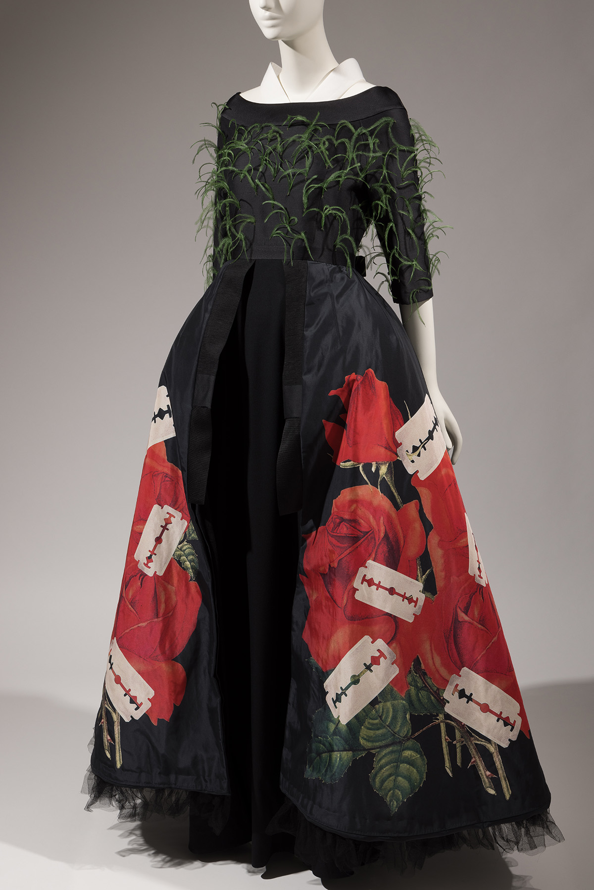 black, 3/4 sleeve dress with green feathers attached to bodice and a skirt with print of red roses and razorblades