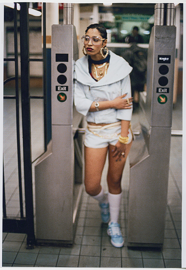 a woman wearing a white streetwear ensemble and gold jewelry posing in a subway station