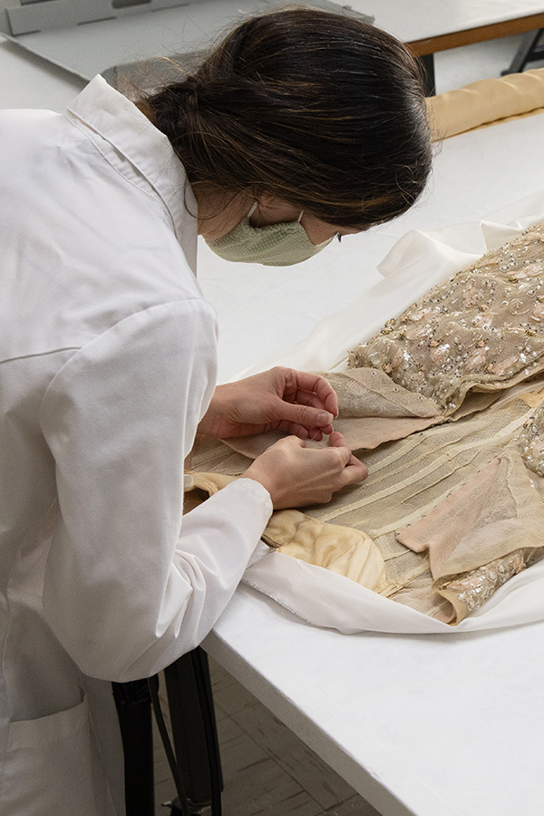 woman in a lab coat and mask hand sewing the back closure of a ballgown in a conservation lab