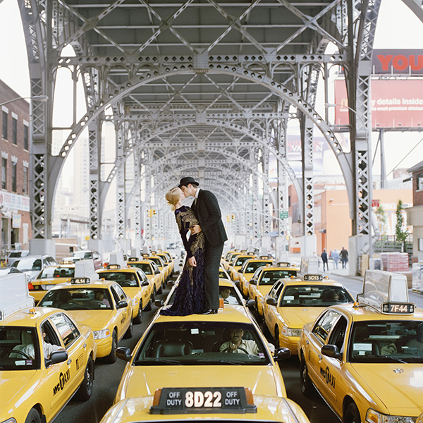 a couple kisses standing on a yellow cab amidst a sea of yellow cabs under an elevated expressway