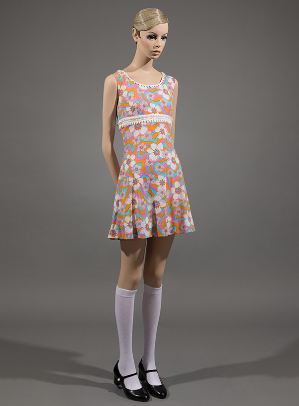 twiggy mannequin wearing sleeveless short length dress with a pink, yellow, blue, and white flower print, styled with white knee high socks and black mary jane shoes