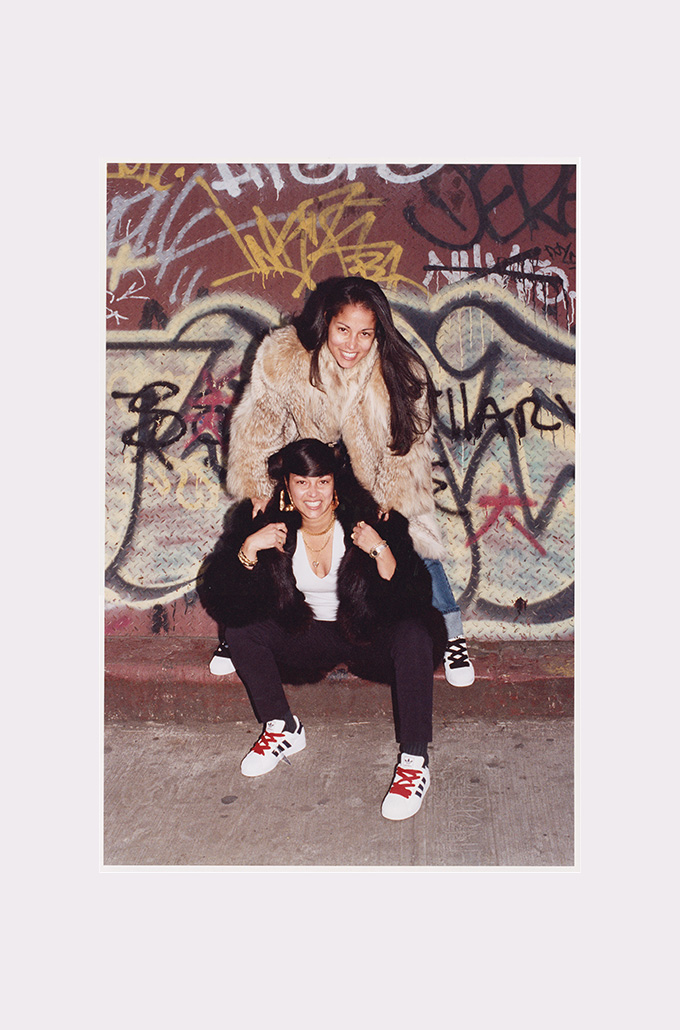 two women smiling and posing on a street against a graffiti wall