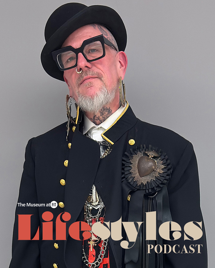 Wren wearing a black bowler hat with black, square rimmed glasses, hoop earrings, a crucifix earring, and a black suit jacket with gold embellishments and a sacred heart in 