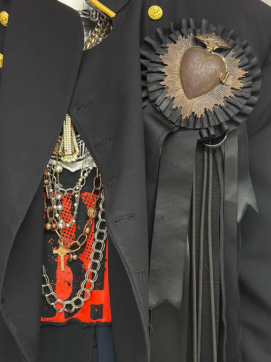 bronze sacred heart pin wrapped in black ribbon on a black suit jacket and chained necklaces