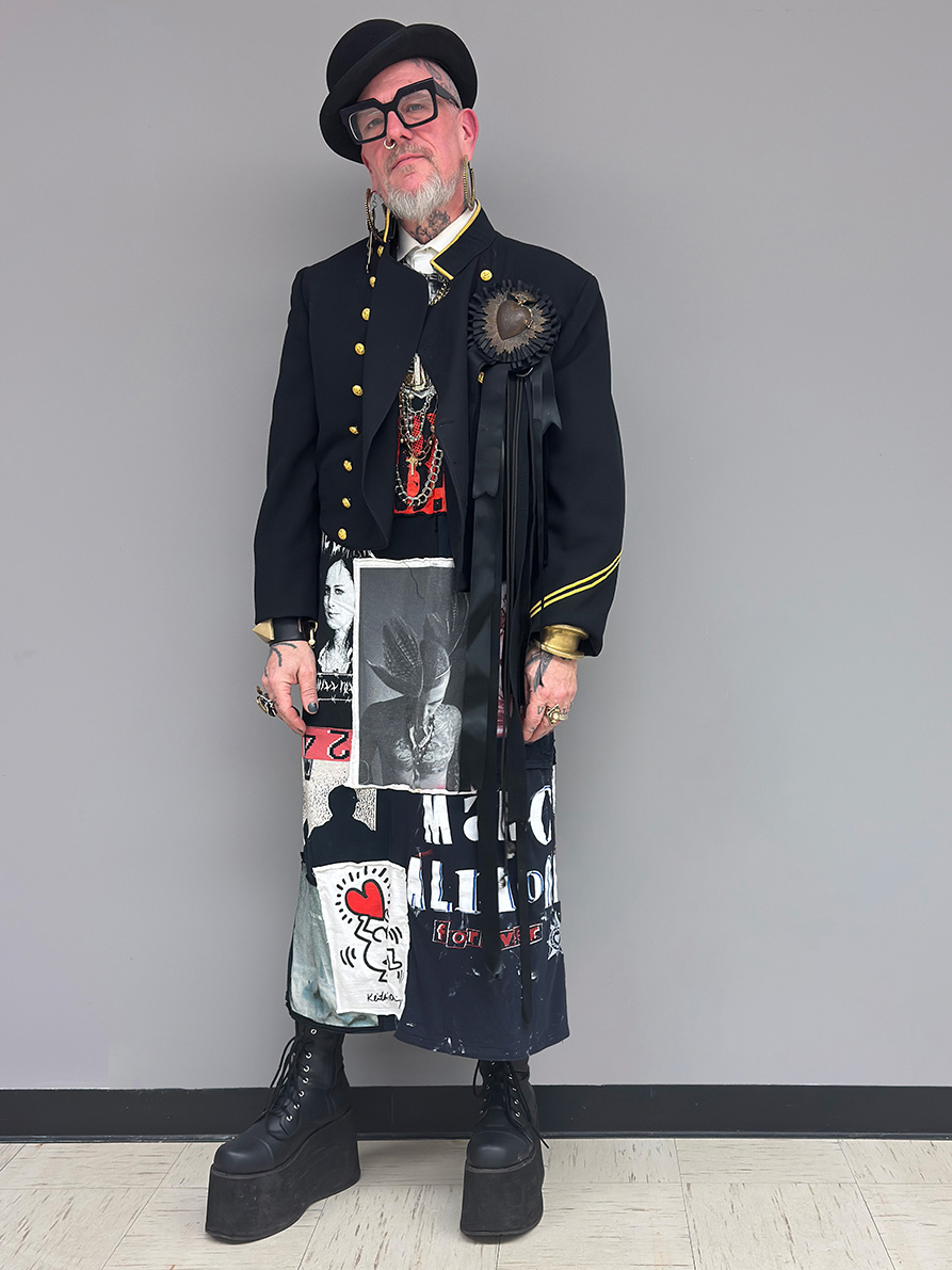 an older white man, Wren Britton, wearing a black suit jacket styled over an ankle length dress of patchwork t-shirts, styled with think black rimmed glasses, a bolwer hat, crucifix earrings, and black platform lace up boots