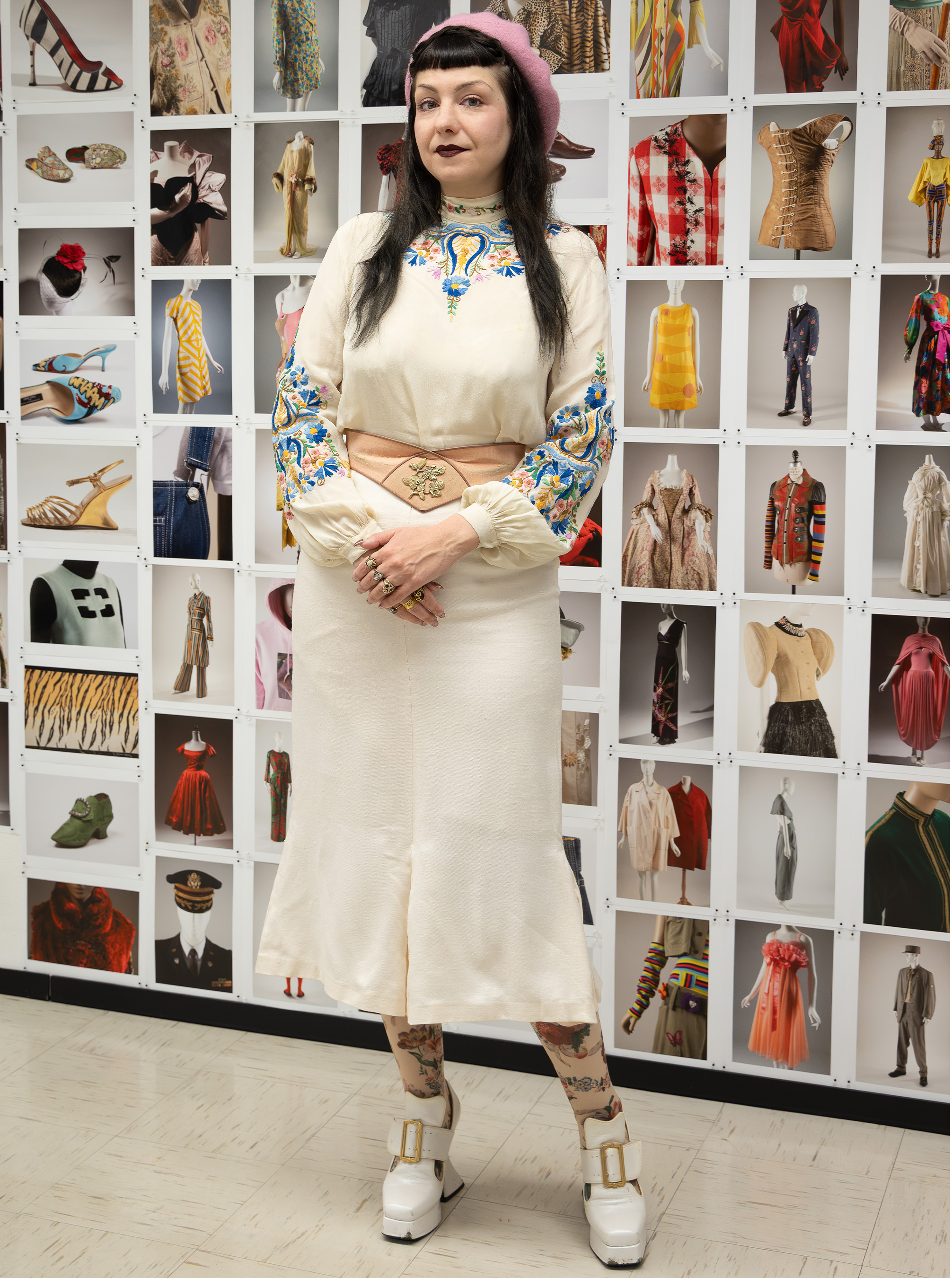 elena standing in front a gallery wall, wearing a white long sleeved embroidered top paired with a white skirt, pink beret, tan belt, and white chunky high heeled shoes