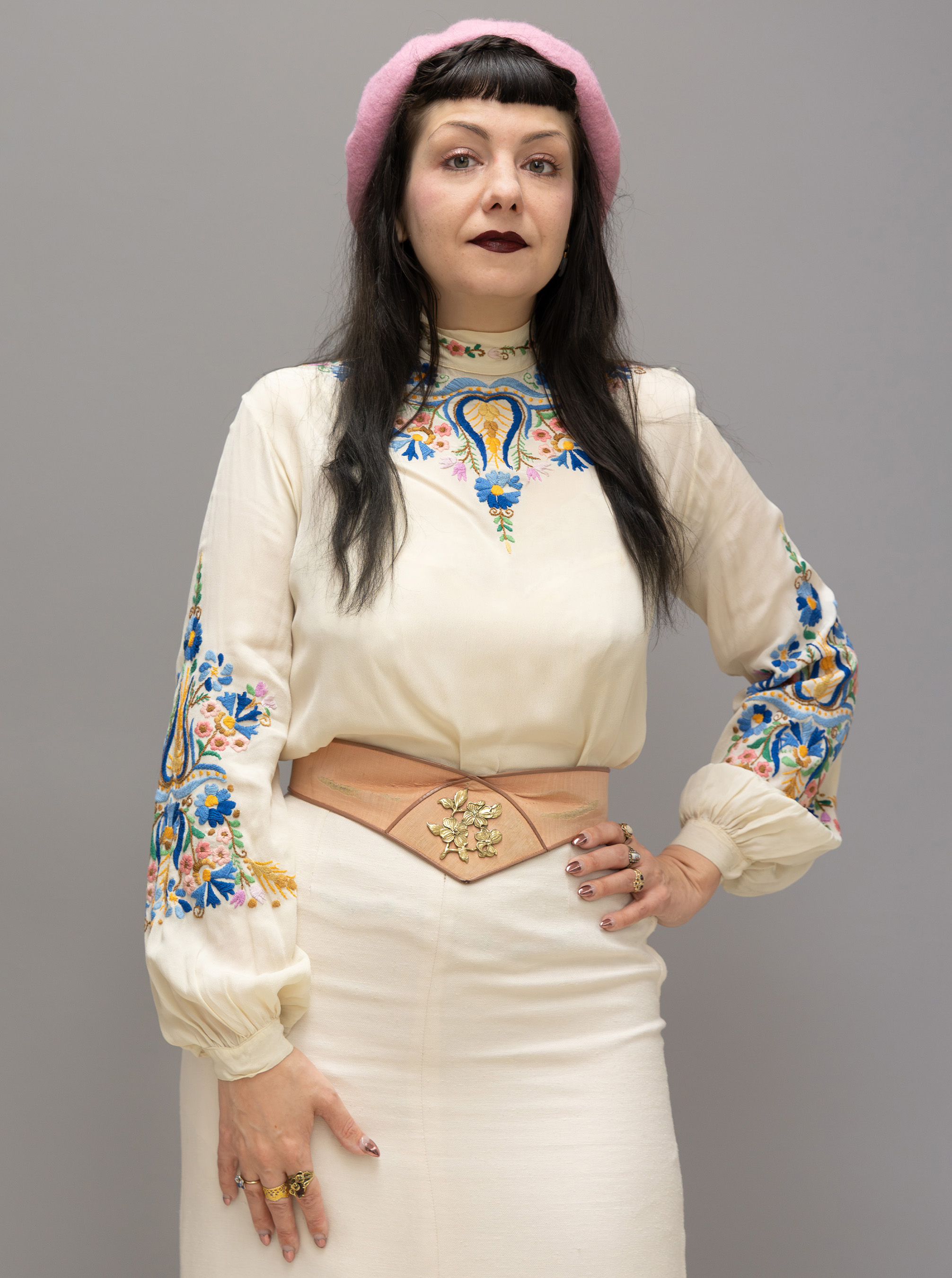 elena standing with a hand on her hip, showing off her embroidered sleeves and tan belt against her white top and skirt