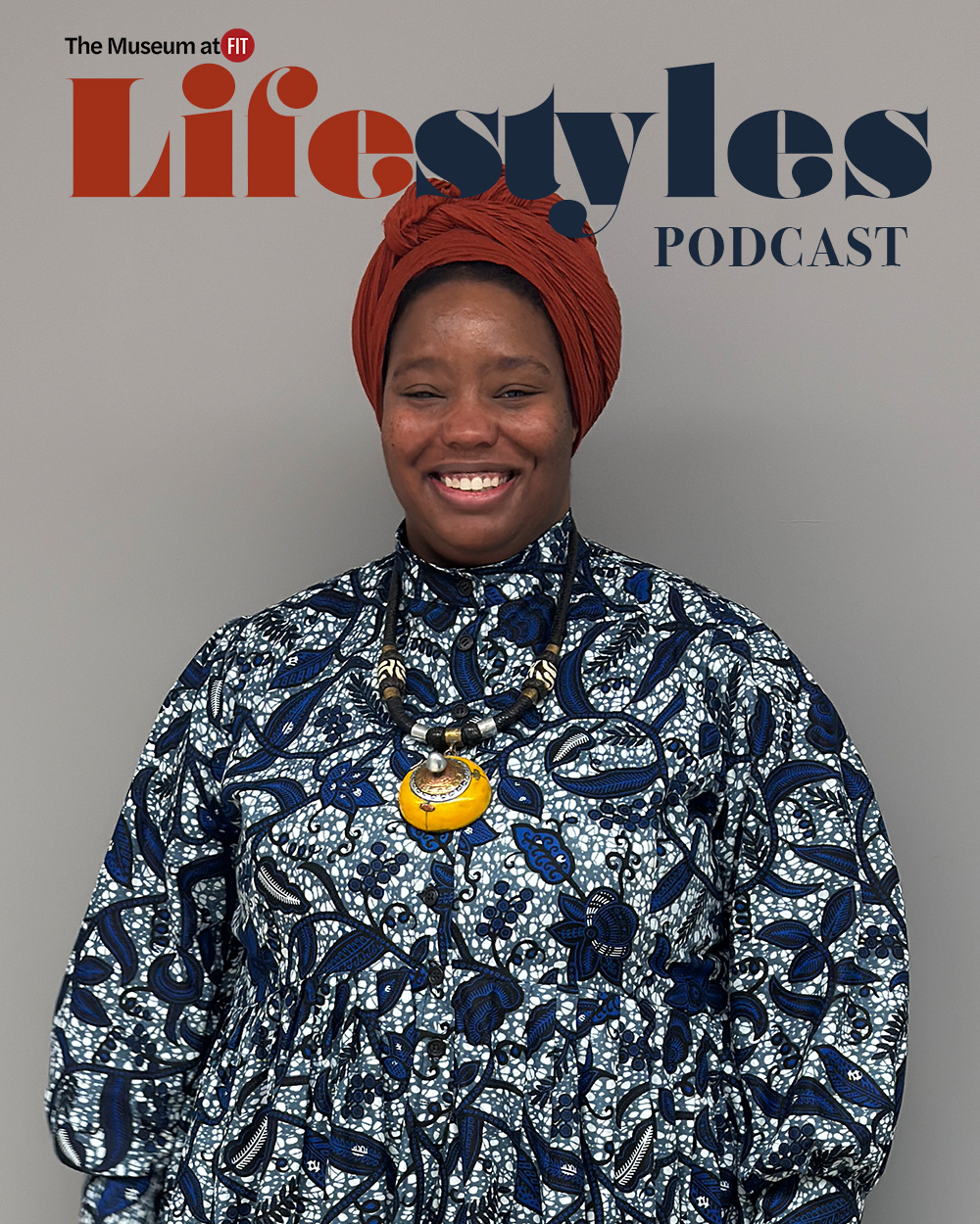 Cheyney smiling and wearing a blue, white, and grey long sleeved button down dress styled with a rust colored head wrap and a black braided necklace featuring a chunky, circle pendant