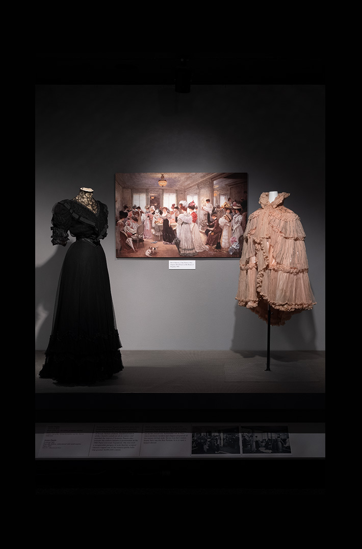 Designing Women installation of a dress and a cape in front of an impressionist painting of women dressed in the style of the object on view 