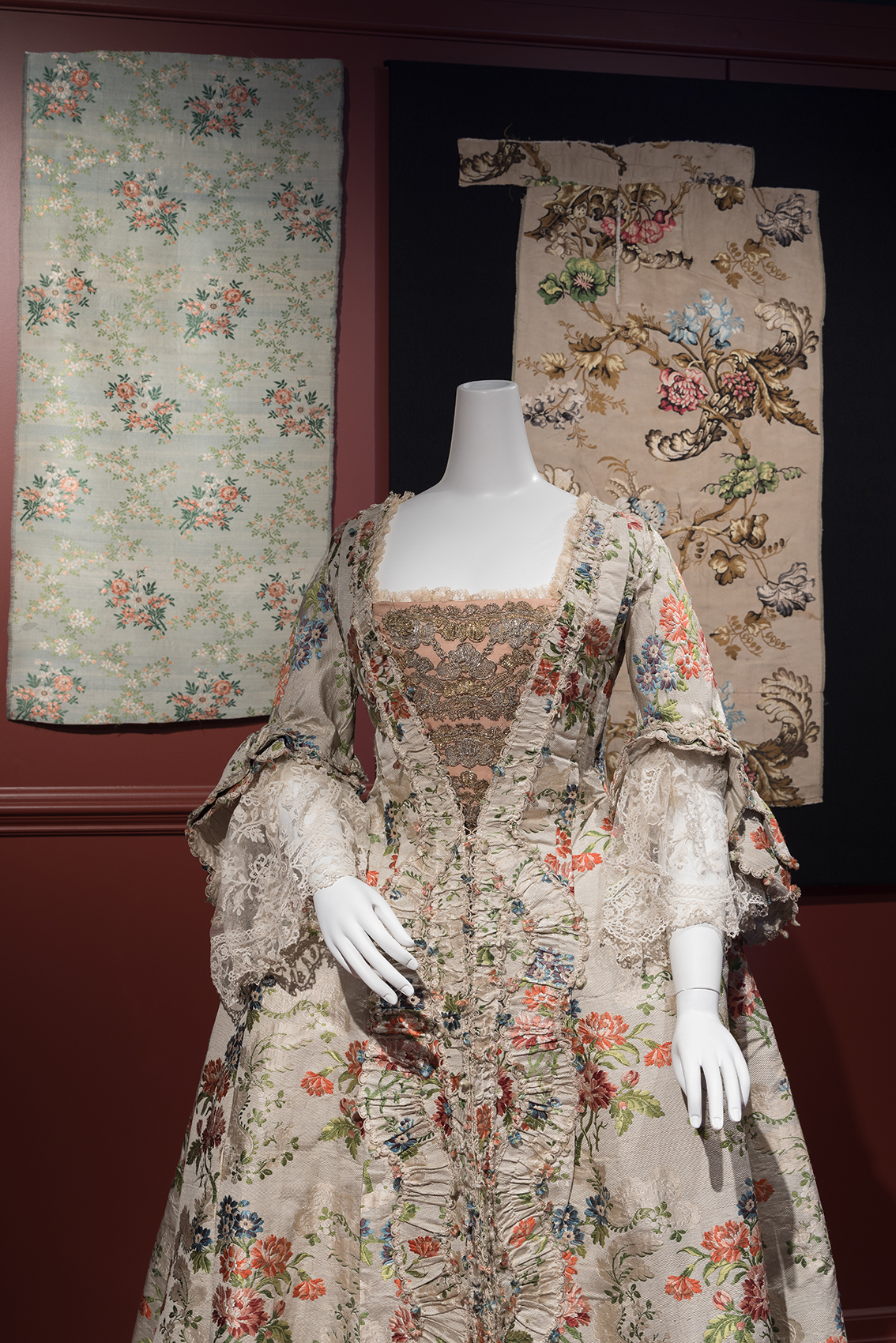 ivory floral brocade robe a la francaise on a mannequin in front of alternative floral fabrics hanging against a dark wall 