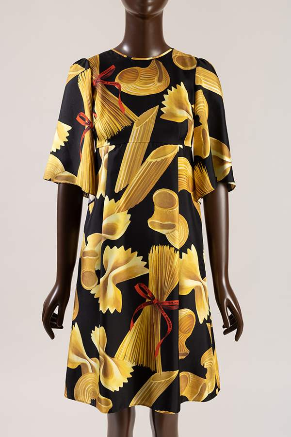 Knee-length silk dress with yellow gold penne, farfalle, macaroni and spaghetti pasta print tied with red logo ribbon on black ground 