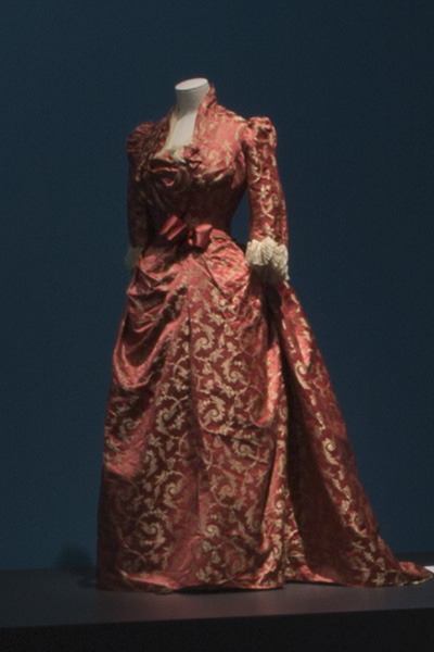 Floorlength red dress with gold pattern, high collar, and rose at bosom.