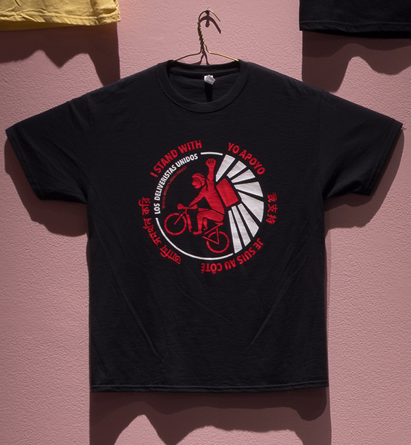 Black t-shirt with an illustrated delivery person on a bike in red surround in a circle by text that reads I stand with you apoyo