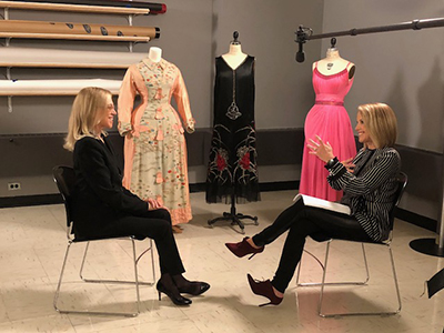 Valerie Steele siting across from Katie Couric with three dress objects in the background