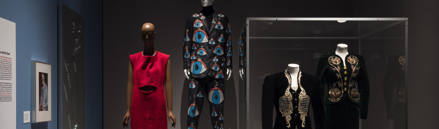 gallery featuring a broken mirror on the left and mannequins wearing a red dress, men's suit ensemble with painted eyes, and two jackets with a mosaic of mirror shards