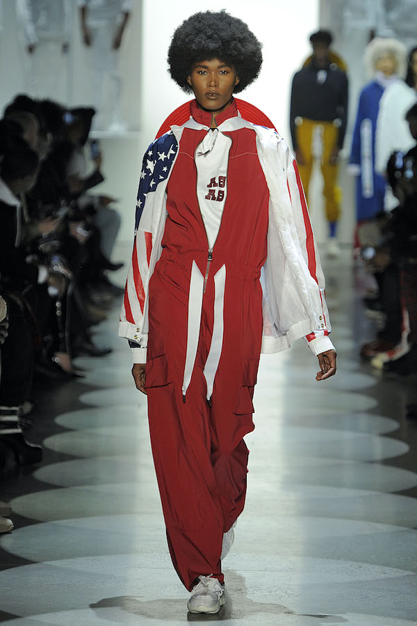 Male model walking a runway in a red jumpsuit with an American flag cape