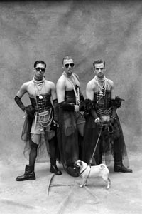 black and white image of three man in black corsets with tulle skirts and boots staring into the camera with one man holding a leash to a pug