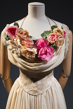 close-up view of cream-colored sleevless dress with scoop neck and corseted waist. A bouquet of silk flowers adorns the bosom.
