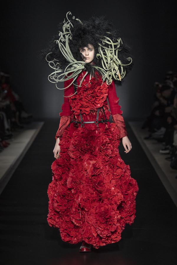 runway image of a model in a full length red dress that has surface ruffles that appear as roses