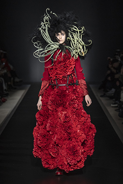 runway image of a model in a full length red dress that has surface ruffles that appear as roses