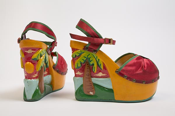 Red ankle strap platform wedge open toe sandals in synthetic satin with hand painted and carved relief design on polyurethane platform base of palm trees and tropical beach scene in red, yellow, blue, green, orange, brown and white, upper front panel cinched at CF and held in place with self fabric band, green fabric bias binding at finished edges, upper shoe attached to base with tarnished gold metal nail heads and leather trim with  fluted copper metal stud at inside strap, copper metal buckle at ankle strap closure