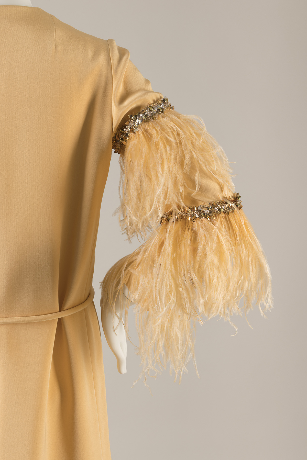 Partial side view of a silk dress sleeve trimmed with feathers and horizontal bands of rhinestones and beads at mid-arm.