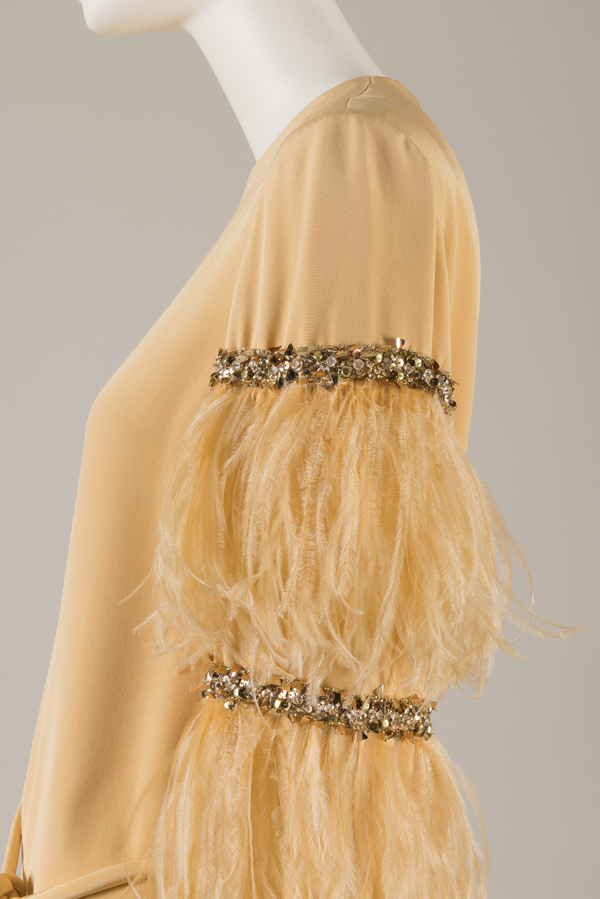 Partial side view of a silk dress sleeve trimmed with feathers and horizontal bands of rhinestones and beads at mid-arm.