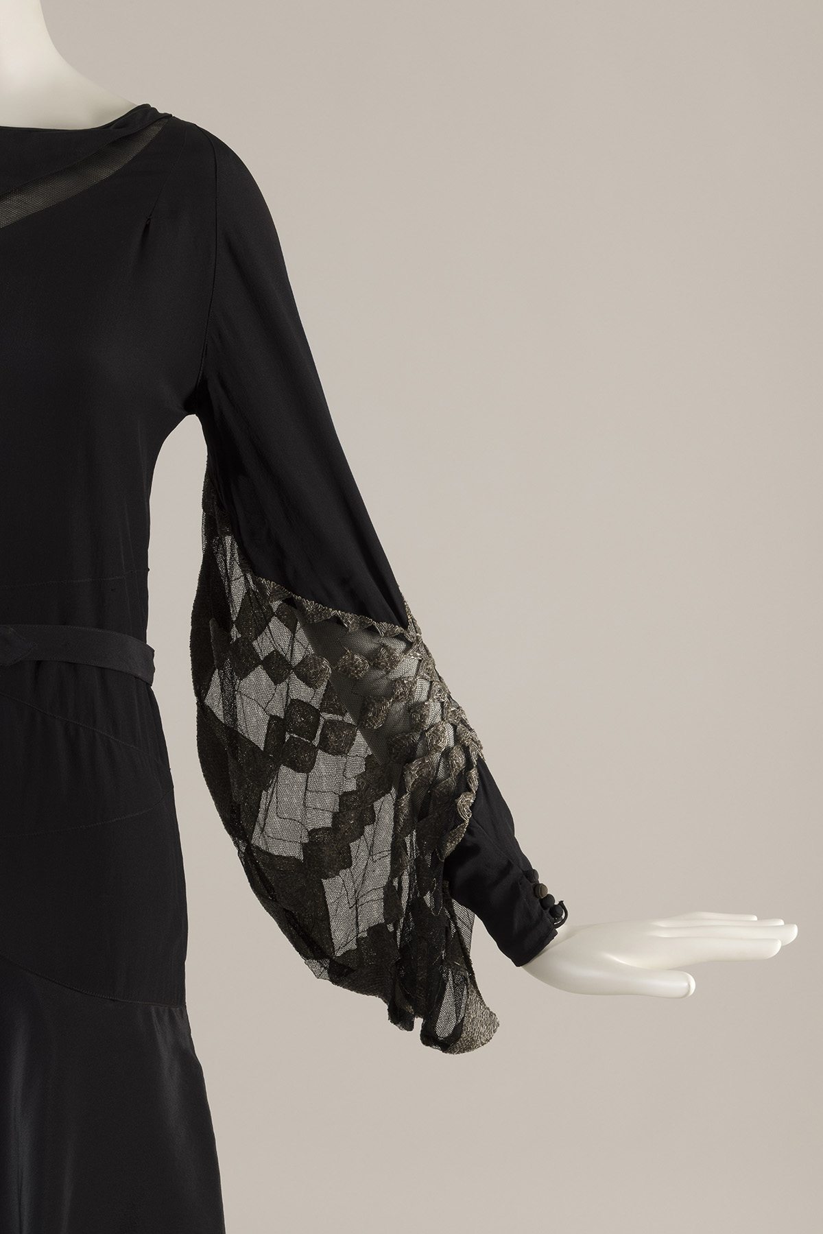 A black silk dress with a straight silhouette. The lower portion of the dress’s long sleeve features an insert of black net embroidered with silver beads in a geometric pattern.
