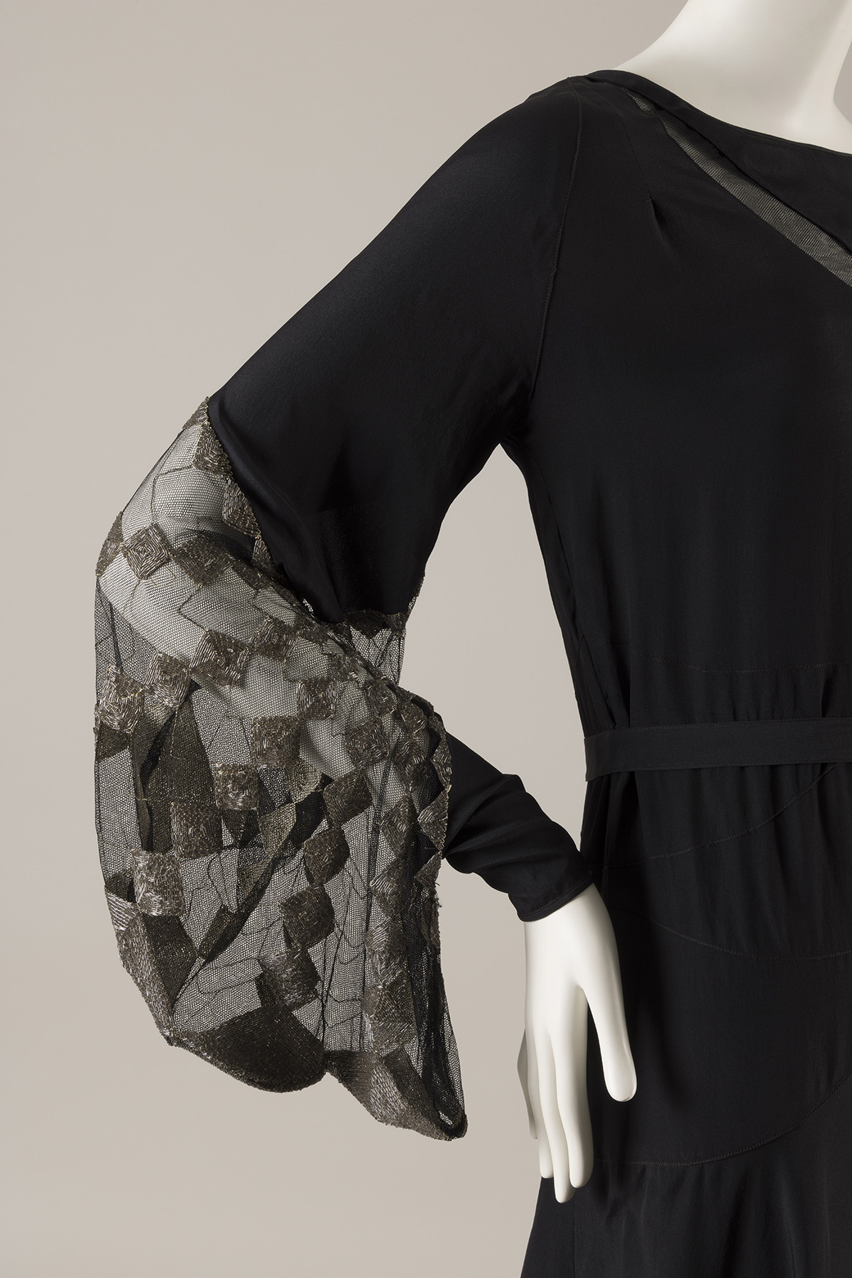 A black silk dress with a straight silhouette. The lower portion of the dress’s long sleeve features an insert of black net embroidered with silver beads in a geometric pattern. 