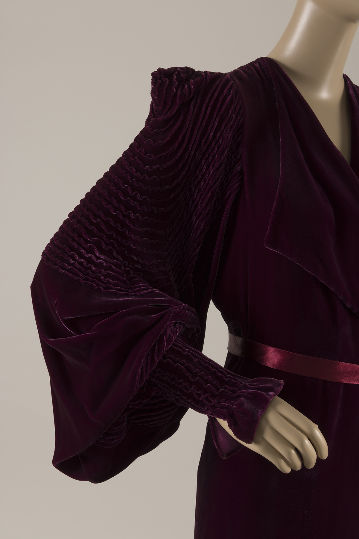 Partial side view of a woman’s dressing gown in velvet, tied at the waist with a matching silk ribbon. The full sleeve has a sharply defined shoulder, a tapered cuff, and elaborate pin-tucks in concentric circles.