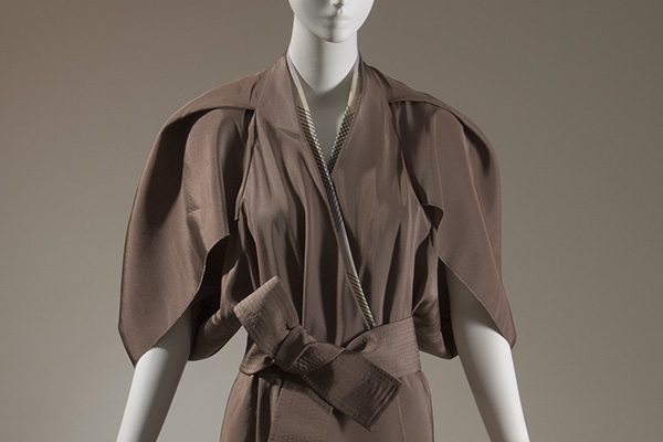 Front view of a dress bodice with fabric that extends like a cape over the shoulders and  gathers into a wide belt at the waist. 