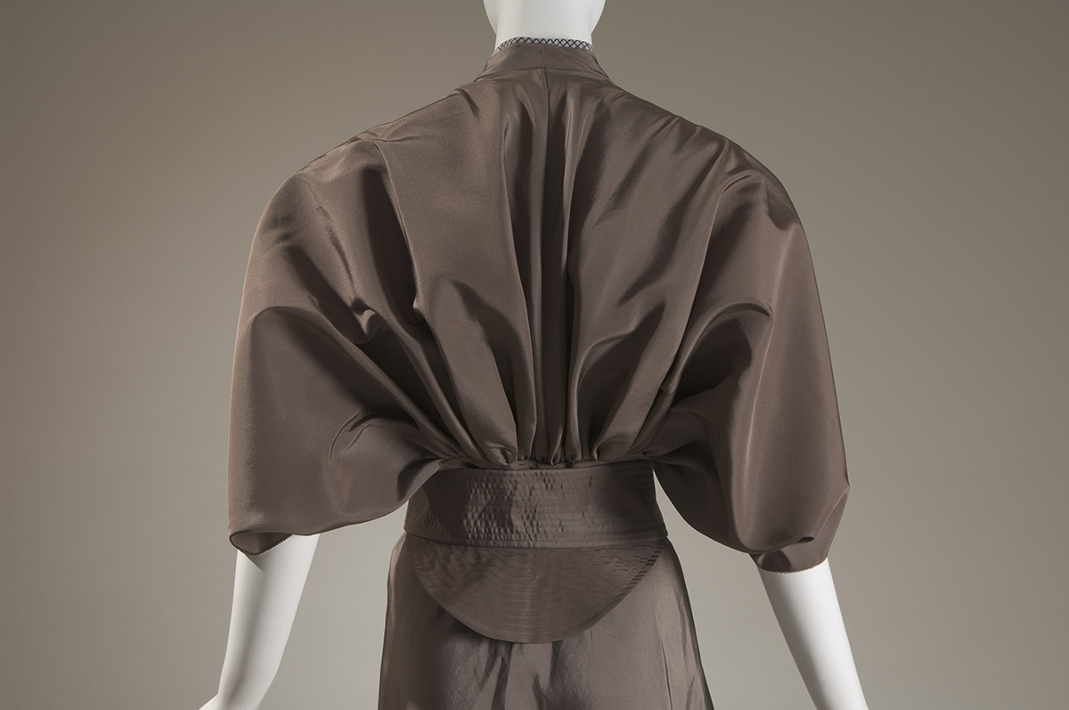 Back view of a dress bodice with fabric that extends like a cape over the shoulders and  gathers into a wide belt at the waist. 