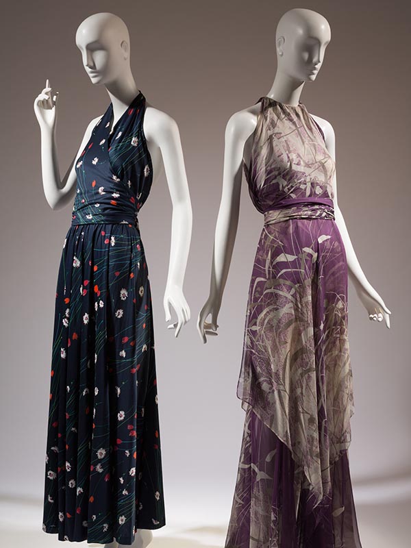two mannequins, the one on the left wearing a navy floor length dress with red, green, and white flower prints and a halter top. The one on the right is wearing a pink silk chiffon floor length dress with a high neckline